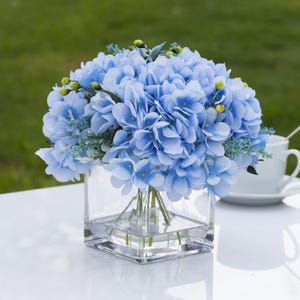 May include: A bouquet of light blue hydrangea flowers in a clear, square glass vase. The arrangement includes small green and yellow accents. The vase sits on a white surface, with a white teacup and saucer in the background.