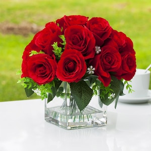 Könnte beinhalten: Ein Strauß leuchtend roter Rosen in einer quadratischen Glasvase. Die Anordnung umfasst grüne Blätter und kleine weiße Blüten, die eine üppige Darstellung schaffen. Die Vase steht auf einer weißen Oberfläche, mit einem verschwommenen grünen Hintergrund.