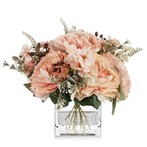 May include: A bouquet of peach-colored peonies and smaller pink flowers with green foliage and dark berries. The arrangement is displayed in a clear, square glass vase, creating a fresh and elegant floral display.