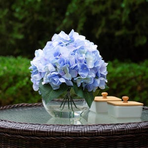 May include: A display of blue hydrangea flowers in a clear glass bowl vase. The flowers are a mix of light and medium blue, with green leaves. Two small white ceramic containers with wooden lids are also present.