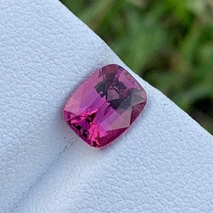 May include: A faceted, rectangular-cut, pink gemstone. The gem is set on a white surface, with a blurred green background. The gemstone's colour ranges from deep magenta to lighter pink hues.