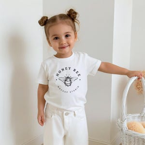 Puede incluir: Un niño pequeño con una camiseta blanca y pantalones blancos. La camiseta presenta un gráfico en blanco y negro de una abeja con el texto "HONEY BEE MOCKUP STUDIO" en un diseño circular. El niño está sonriendo.