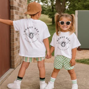 Pode incluir: Duas crianças vestindo camisetas brancas com um design "Honey Bee Mockup Studio" e um gráfico de abelha. Uma criança usa shorts listrados, a outra xadrez. Ambas usam tênis brancos e meias. Uma criança usa um boné marrom e a outra óculos de sol.