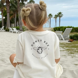 May include: Cream-colored t-shirt with a black and white bee graphic and the text "HONEY BEE MOCKUP STUDIO." The shirt is worn by a child with blonde hair styled in two buns. The background includes palm trees and a beach.