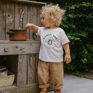 May include: A young child wearing a white t-shirt with a "Honey Bee Mockup Studio" graphic, paired with tan pants and sandals. The child is interacting with a brown pot on a wooden structure, with a green background.