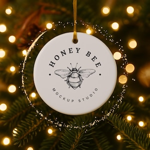 May include: A round, white ceramic ornament with a gold ribbon for hanging. The ornament features a detailed illustration of a bee and the text "HONEY BEE MOCKUP STUDIO" in black. The background is a blurred Christmas tree with warm lights.