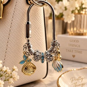 May include: A silver-toned metal carabiner bag charm featuring a central bow-shaped bead encrusted with clear and blue crystals, flanked by two silver beads, a gold-toned charm with blue enamel, and a gold-toned seashell charm with blue enamel.
