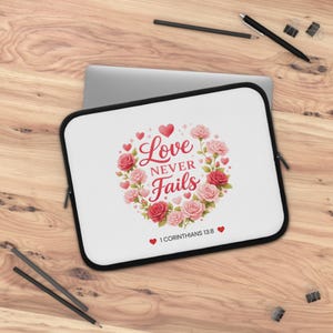Puede incluir: Funda blanca para portátil con borde negro, con un diseño floral de corazón con rosas rojas y rosas. El texto "Love Never Fails" está centrado, con "1 Corintios 13:8" debajo.