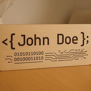 May include: A rectangular light-colored wooden sign featuring dark engraved text that reads <{ John Doe }; followed by binary code 01010110100 00100011010 and a graphic circuit board pattern. The sign has rounded corners and a smooth finish.