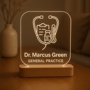 Puede incluir: Un letrero acrílico iluminado con un diseño de estetoscopio, historial médico y frasco de medicina. El letrero está personalizado con el texto "Dr. Marcus Green" y "GENERAL PRACTICE" en blanco. Se encuentra sobre una base de madera.