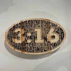 Puede incluir: Placa ovalada de madera con texto tallado. El texto es de color oscuro y dice: "Porque de tal manera amó Dios al mundo, que ha dado a su Hijo unigénito, para que todo aquel que en él cree, no se pierda, mas tenga vida eterna."