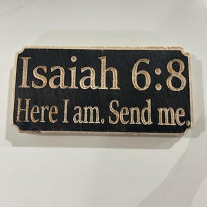 Puede incluir: Un letrero rectangular de madera con el texto "Isaiah 6:8 Here I am. Send me." El letrero tiene un fondo negro con el texto en color marrón claro. Los bordes del letrero son redondeados.