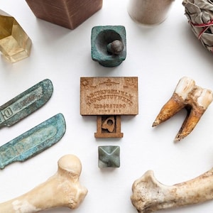 May include: An assortment of spiritual and occult items, including a wooden Ouija board, crystals, bone fragments, and aged metal tools. The items are arranged on a white surface, creating a mystical aesthetic.