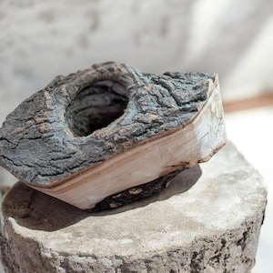 May include: A handcrafted, rustic wooden candle holder. The holder is made from a section of a tree branch, featuring a natural bark exterior in shades of gray and brown. The interior is a smooth, light-colored wood, with a circular opening.