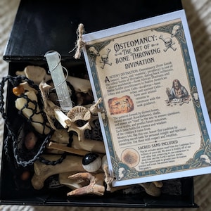 May include: A black box containing a collection of animal bones, a selenite crystal wrapped in wire, a black cord necklace with wooden beads, and a small sphere. An instructional card titled Osteomancy: The Art of Bone Throwing Divination is attached.