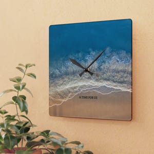 May include: A square wall clock featuring an aerial view of a sandy beach with blue ocean waves crashing against the shore. The clock face displays the text A TIME FOR US in black lettering and includes two black clock hands centered on the image.