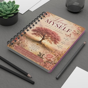 Puede incluir: Un diario de espiral con una cubierta rosa y dorada que presenta un gran árbol, rosas y una mariposa. El texto dice I love MYSELF JOURNAL POSITIVE AFFIRMATIONS, I am beautiful, I am blessed, I choose happiness y Love yourself Heal Grow Glow.