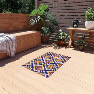 Puede incluir: Una alfombra rectangular para exteriores con un patrón geométrico en tonos azules, amarillos, rojos y blancos. La alfombra está colocada sobre una terraza de madera, con un banco de madera y plantas en el fondo.