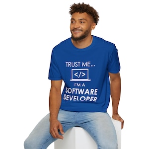 Op de afbeelding: Een koningsblauw t-shirt met de tekst "TRUST ME... I'M A SOFTWARE DEVELOPER" in het wit. Het ontwerp bevat een laptop-pictogram met codesymbolen. De persoon draagt een lichtblauwe jeans.