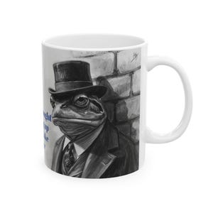 Puede incluir: Taza de cerámica blanca con una ilustración en escala de grises de una rana con traje y sombrero de copa, apoyada en una pared de ladrillos. La obra de arte incluye una lágrima en la cara de la rana. Texto visible en la taza.