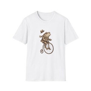 Könnte beinhalten: Weißes T-Shirt mit einer Illustration eines Frosches auf einem alten Fahrrad. Der Frosch trägt einen Anzug, einen Schal und einen Hut. Das Fahrrad ist braun und der Frosch ist beige. Der Hut schwebt über dem Kopf des Frosches.