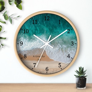 May include: Circular wall clock with a light wood frame. The clock face depicts a beach scene with turquoise water and white waves. The text "A time for us" is printed on the sand. White hands show the time.