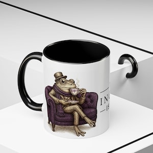 May include: White ceramic mug with a black interior and handle. The mug features a detailed illustration of a frog in a suit and hat, seated on a purple armchair, sipping tea. The text "IN -IS" is partially visible.