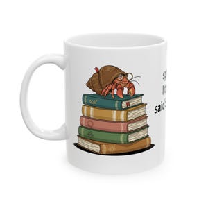 May include: White ceramic mug featuring a cartoon hermit crab wearing glasses and a shell hat, perched atop a stack of colorful books. The mug has a curved handle and the text "sp" and "said" on the side.