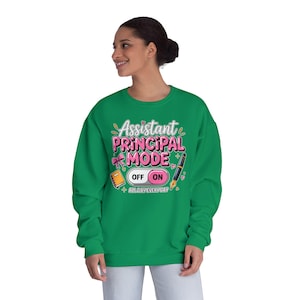 May include: A green sweatshirt with the text "Assistant Principal Mode" in pink and white, with a graphic of a book, a pen, and an on/off switch. The phrase "All Day Every Day" is also included.