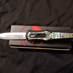 Puede incluir: Un cuchillo automático negro y plateado con mango de concha de abulón descansa sobre su caja original. La hoja es plateada con borde dentado y el mango tiene un marco negro. La caja es negra con un diseño circular rojo.