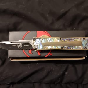 May include: A tactical knife with a silver blade and a handle inlaid with iridescent abalone shell. The knife rests on a black box with red accents. The blade has the text "2794" and "10/2004".