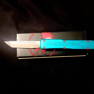 Può includere: Un coltello automatico turchese e argento con un accento rosso, appoggiato su una scatola nera con un logo rosso. Il coltello ha una lama argentata e un manico turchese con dettagli rossi. Lo sfondo è nero.