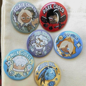 Puede incluir: Un conjunto de seis botones redondos con diseños de personajes de dibujos animados. Cada botón presenta un nombre de personaje diferente, como "Eclair Cookie" y "Dark Choco". Los botones son de colores brillantes y tienen un acabado brillante.