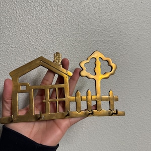 May include: A brass key holder featuring a house, tree, and picket fence design. The key holder has multiple hooks for keys and is made of a gold-colored metal. The house has a chimney and windows.