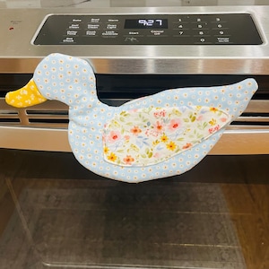 Peut inclure: Un gant de cuisine en forme de canard. Le gant est bleu clair avec de petits motifs floraux blancs et jaunes. Le centre du gant présente un motif floral avec des fleurs roses, jaunes et blanches. Le bec du canard est jaune.