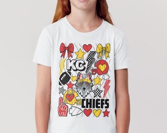 May include: White t-shirt with a colourful graphic design including the text "KC Chiefs". The design features hearts, stars, bows, and a wolf wearing sunglasses. The shirt is worn by a person.