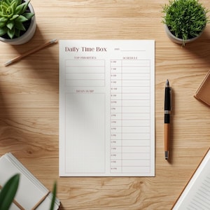 Könnte beinhalten: Ein weißer Daily Time Box-Planer mit einem Zeitplan und einem Bereich für Top-Prioritäten. Der Planer, mit einem Brain-Dump-Bereich, liegt auf einer Holzoberfläche. Ein Stift und kleine Topfpflanzen sind ebenfalls zu sehen.