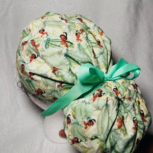 May include: A green and white patterned scrub cap featuring a repeating design of a princess in a green dress. The cap is tied with a light green ribbon. The cap is designed to be worn in a medical setting.