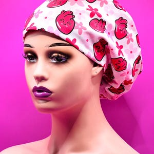 May include: A white scrub cap with a pink heart and flower pattern. The cap is made of a soft fabric and is designed to be worn in a medical setting. The cap is shown on a mannequin with a pink background.