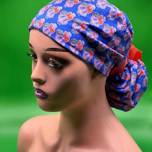 May include: A blue patterned scrub cap with a red ribbon. The cap features a repeating design of white and orange floral shapes on a blue background. The cap is worn by a mannequin with a green background.