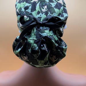 May include: A patterned scrub cap featuring a black bow. The cap is decorated with a repeating design of cartoon characters, hearts, and the words "NOT YOURS" in green and black.