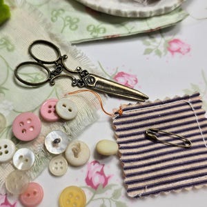 May include: A collection of sewing supplies including a pair of ornate scissors, various buttons in shades of pink, white, and yellow, a striped fabric swatch with a safety pin, and spools of thread. The items are arranged on a floral patterned surface.