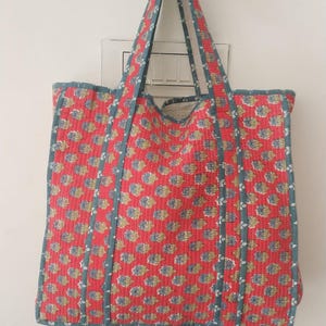 May include: A quilted tote bag with a red floral pattern and blue trim. The bag has two long handles and is hanging against a white wall. The bag's dimensions are not visible.