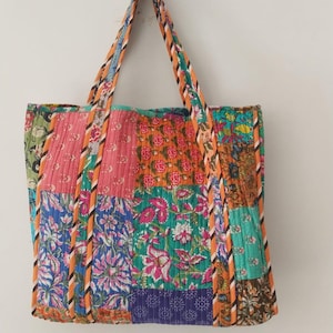 May include: A patchwork tote bag with a variety of floral patterns in pink, blue, green, and orange. The bag has matching handles and trim. The bag is hanging on a white wall.