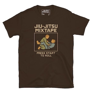 May include: A brown t-shirt featuring the text "Jiu-Jitsu Mixtape" above a pixelated graphic of two people grappling. Below the graphic is the text "Press Start to Roll". The shirt has a crew neck and short sleeves.