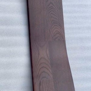 K&ouml;nnte beinhalten: Ein gebogenes, dunkelbraunes Holzbrett mit abgerundeter Oberkante. Die Holzmaserung ist sichtbar und erzeugt eine strukturierte Oberfl&auml;che. Das Brett scheint ein Bestandteil eines gr&ouml;&szlig;eren M&ouml;belst&uuml;cks zu sein, m&ouml;glicherweise eine Stuhllehne oder eine Tischkante.