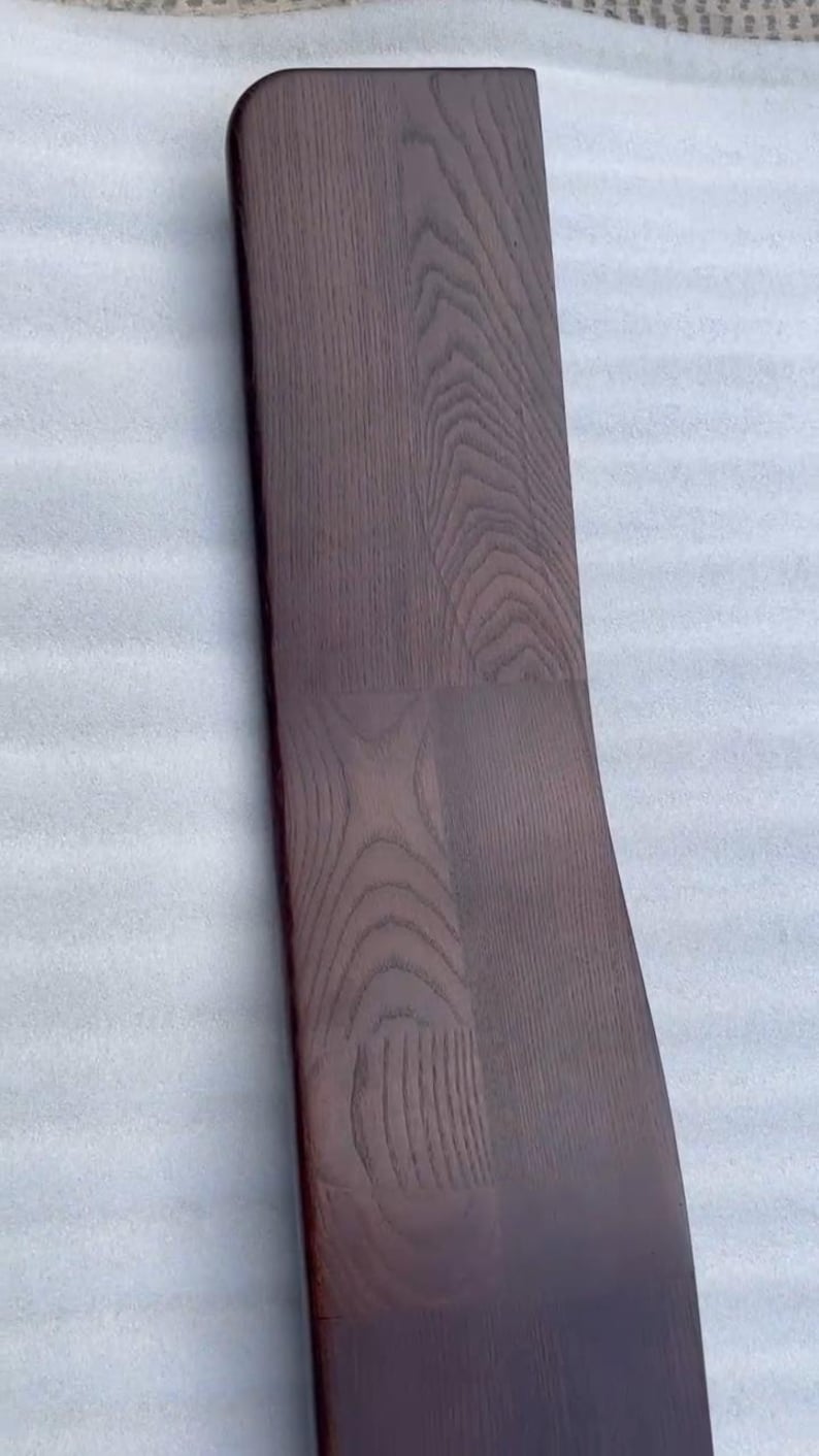 K&ouml;nnte beinhalten: Ein gebogenes, dunkelbraunes Holzbrett mit abgerundeter Oberkante. Die Holzmaserung ist sichtbar und erzeugt eine strukturierte Oberfl&auml;che. Das Brett scheint ein Bestandteil eines gr&ouml;&szlig;eren M&ouml;belst&uuml;cks zu sein, m&ouml;glicherweise eine Stuhllehne oder eine Tischkante.