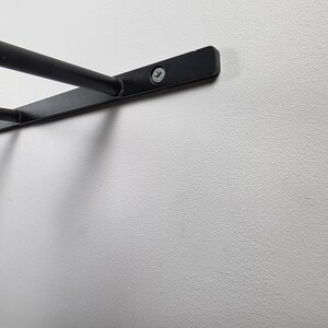 K&ouml;nnte beinhalten: Nahaufnahme einer schwarzen, an der Wand befestigten Regalhalterung aus Metall. Die Halterung hat ein gebogenes Design mit zwei horizontalen Stangen und ist mit Schrauben an einer wei&szlig;en Wand befestigt. Das Regal ist f&uuml;r die Aufbewahrung oder Pr&auml;sentation konzipiert.