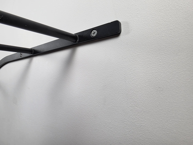 K&ouml;nnte beinhalten: Nahaufnahme einer schwarzen, an der Wand befestigten Regalhalterung aus Metall. Die Halterung hat ein gebogenes Design mit zwei horizontalen Stangen und ist mit Schrauben an einer wei&szlig;en Wand befestigt. Das Regal ist f&uuml;r die Aufbewahrung oder Pr&auml;sentation konzipiert.