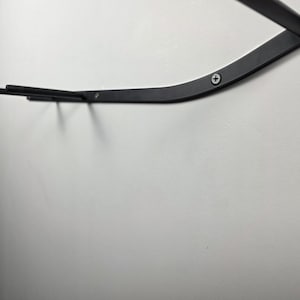 K&ouml;nnte beinhalten: Ein schwarzes Metall-Wandregal mit geschwungenem Design. Das Regal ist an einer wei&szlig;en Wand befestigt und mit Schrauben gesichert. Das Regal hat zwei horizontale Stangen, die sich von der Hauptst&uuml;tze erstrecken.
